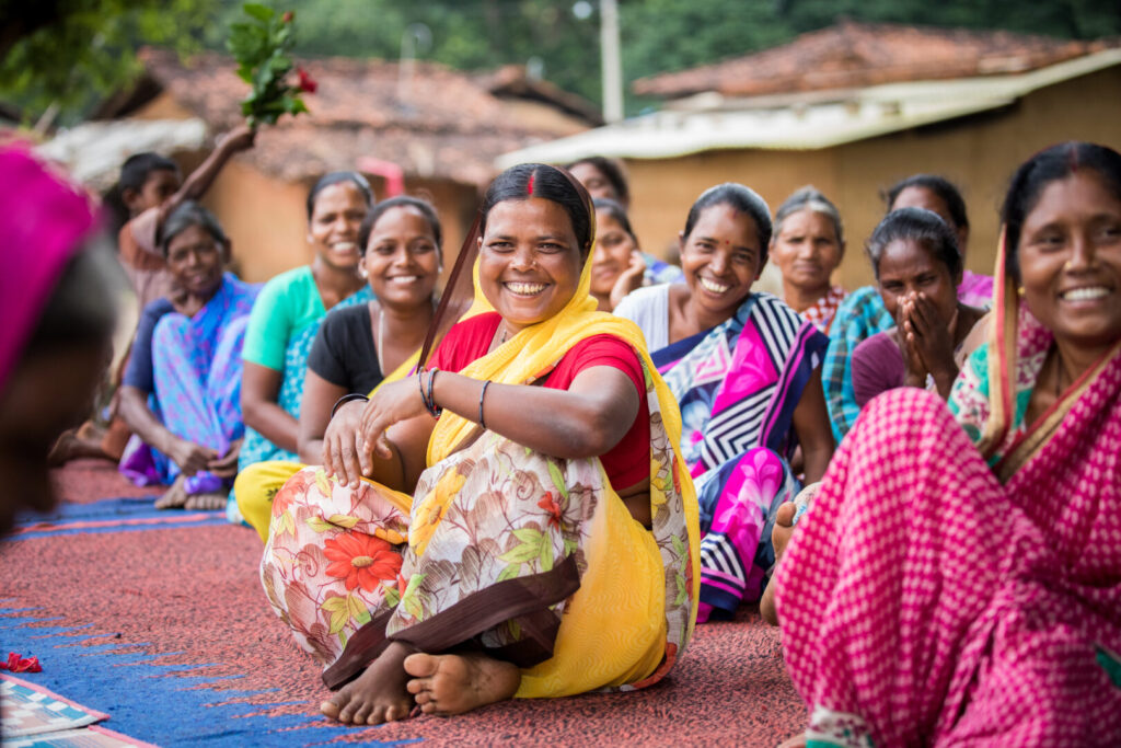 Women in colorful Saris smiling and laughing while sitting on a rug outside.